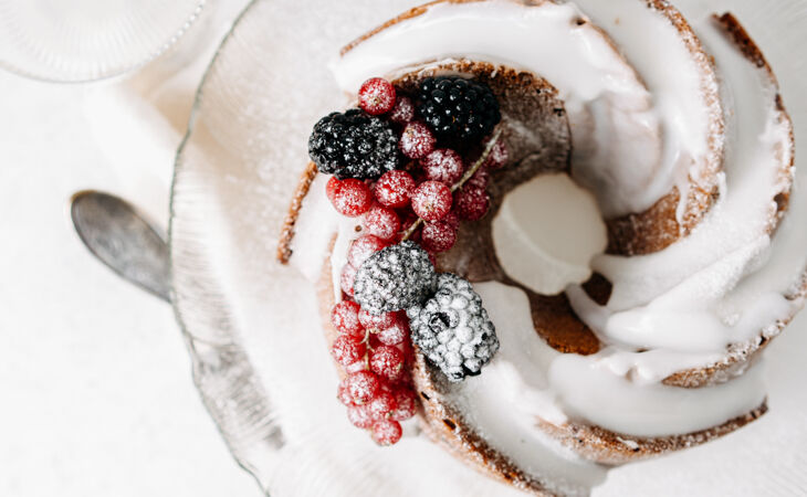 A glazed birthday bundt cake topped with berries and dusted with icing sugar, presented on a glass plate.